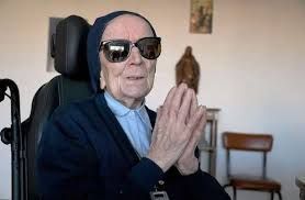 An elderly woman wearing sunglasses is sitting in a wheelchair.