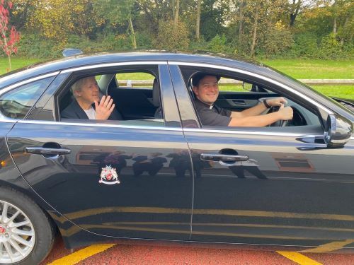 Two men are sitting in a black car waving at the camera.
