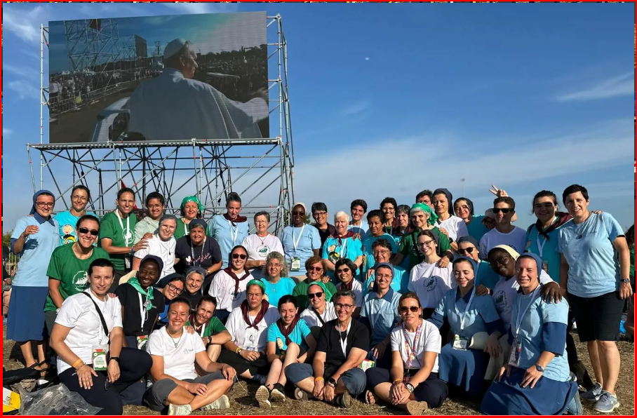 Group of people in blue and green pose in front of a screen showing an event, outdoors under a clear sky.