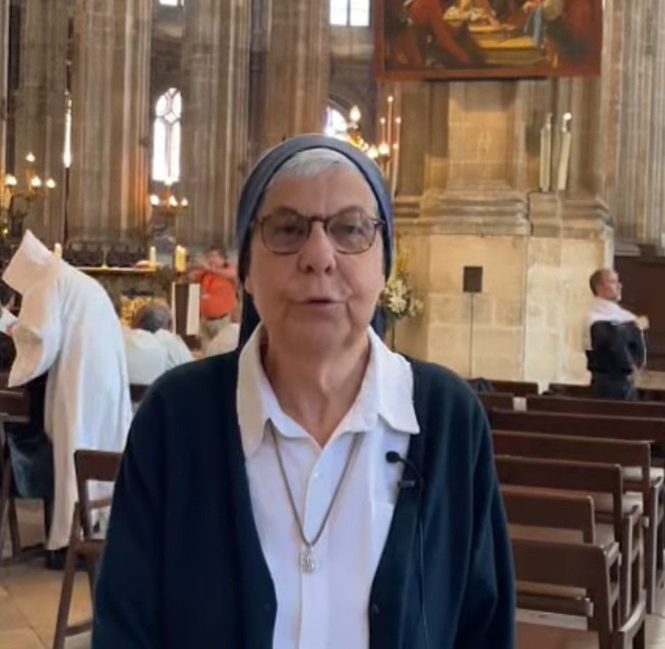 Nun in church, wearing glasses, speaking. People in the background.