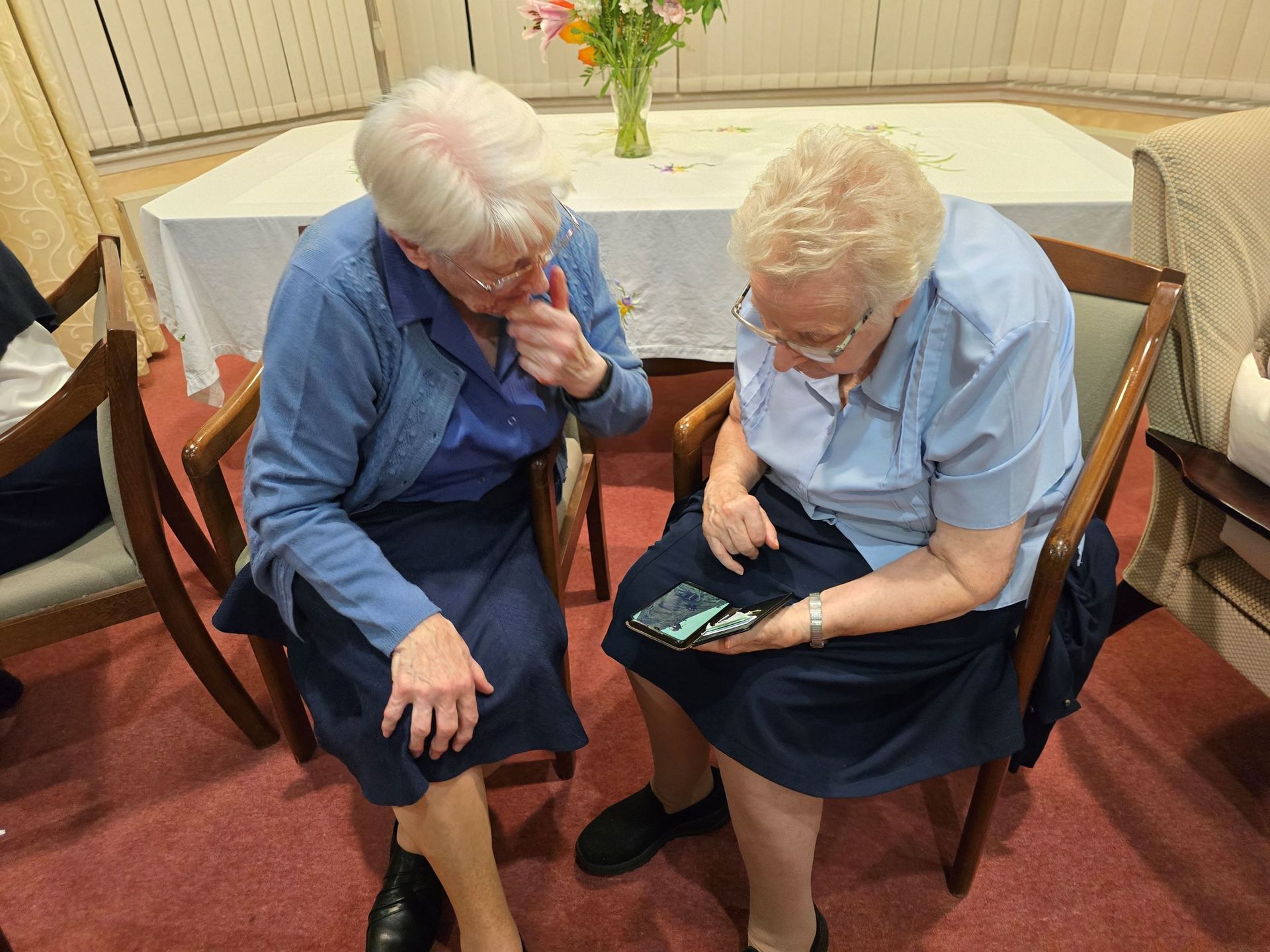 Two older women are sitting in chairs looking at a cell phone.