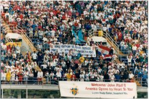 A large crowd of people in a stadium with a banner that says welcome john paul ii america opens its heart to you