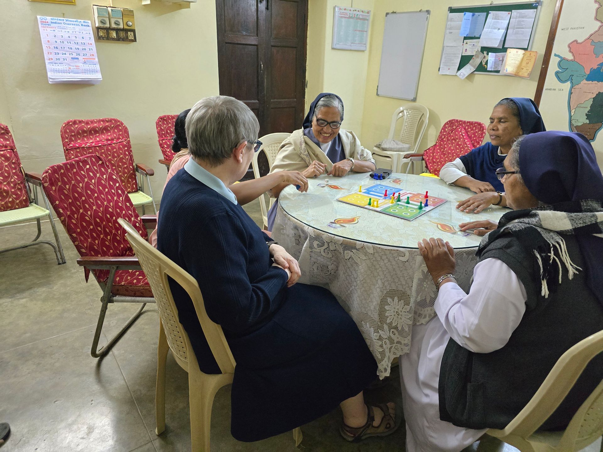 Five nuns play a board game around a table in a brightly lit room.