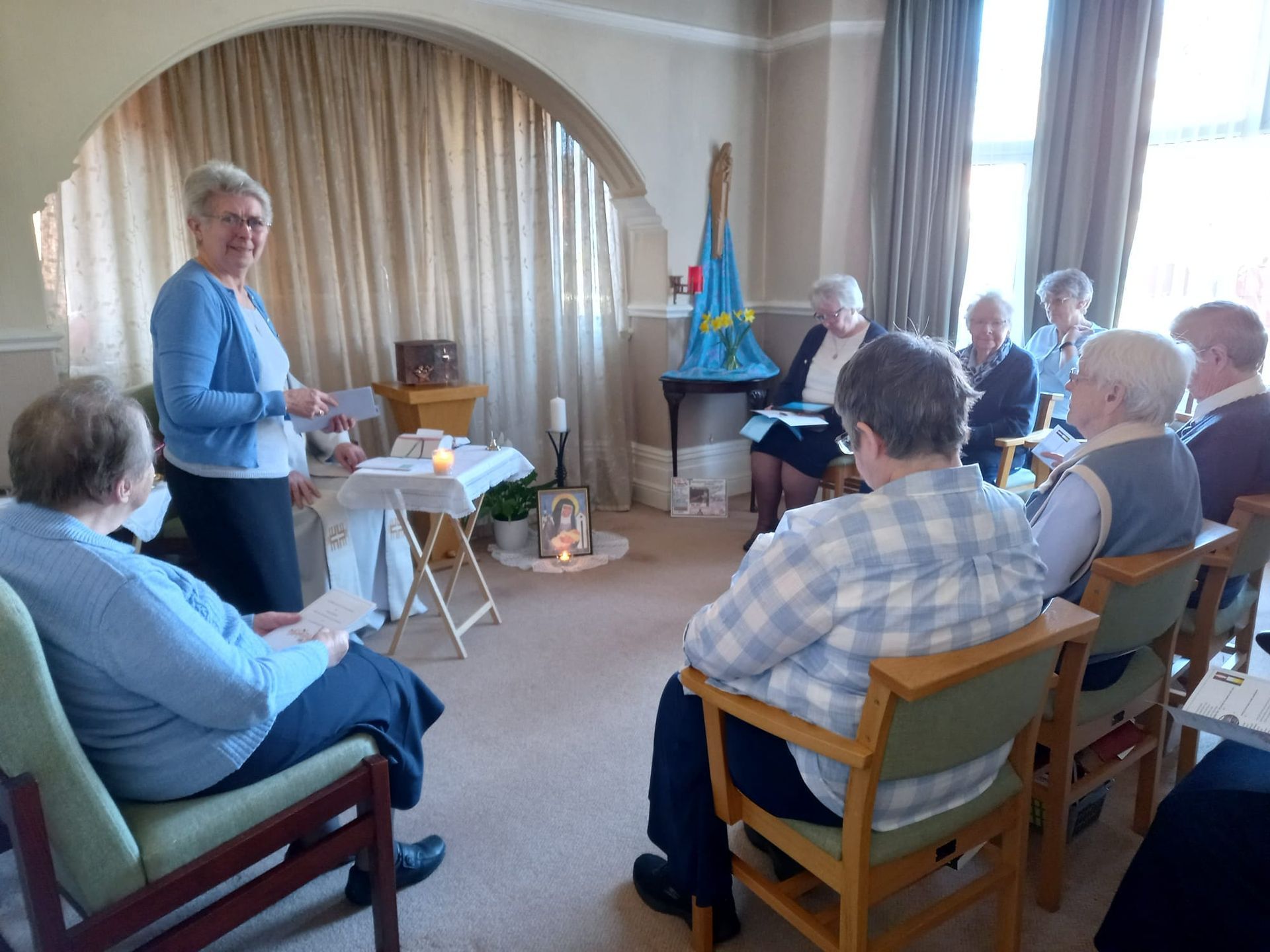 Group of people in a room listening to a woman speaking. Candles, altar, and a blue and yellow decoration are present.