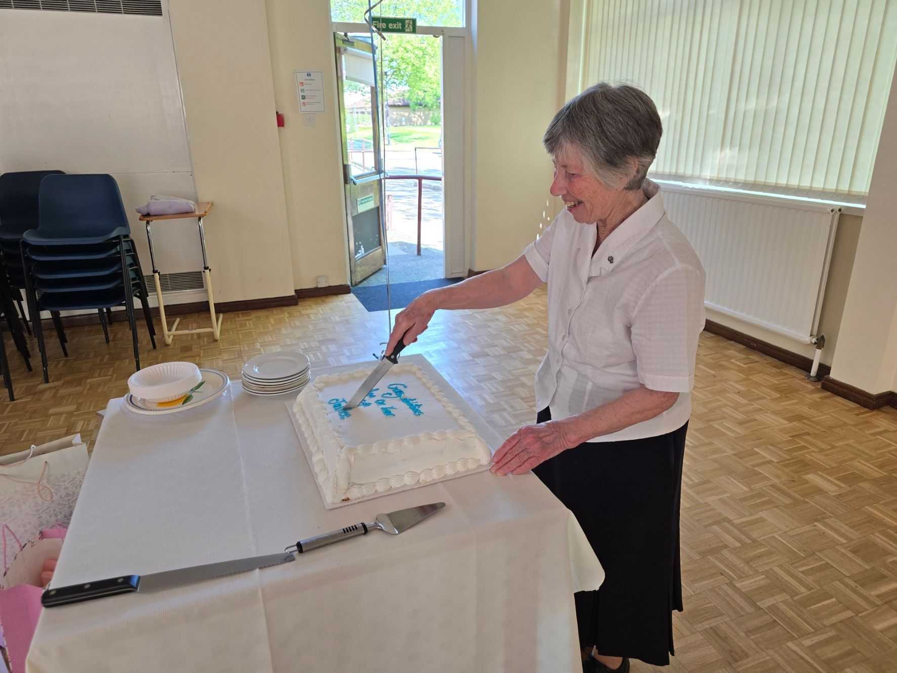 Woman cutting a white cake with blue writing at a table in a brightly lit room.