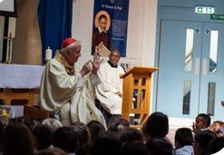 Cardinal speaking to a seated crowd. Other religious figures near a lectern. Banner of a saint visible.