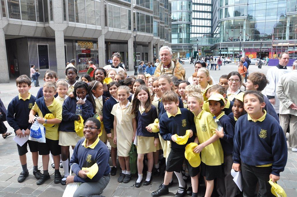 Schoolchildren posing outside, some holding yellow hats, near modern buildings.