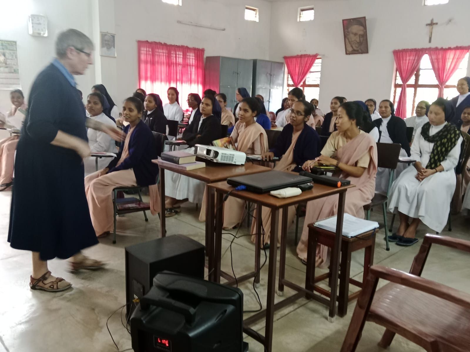 A woman lectures to a group of women in a classroom. Most are wearing religious habits.
