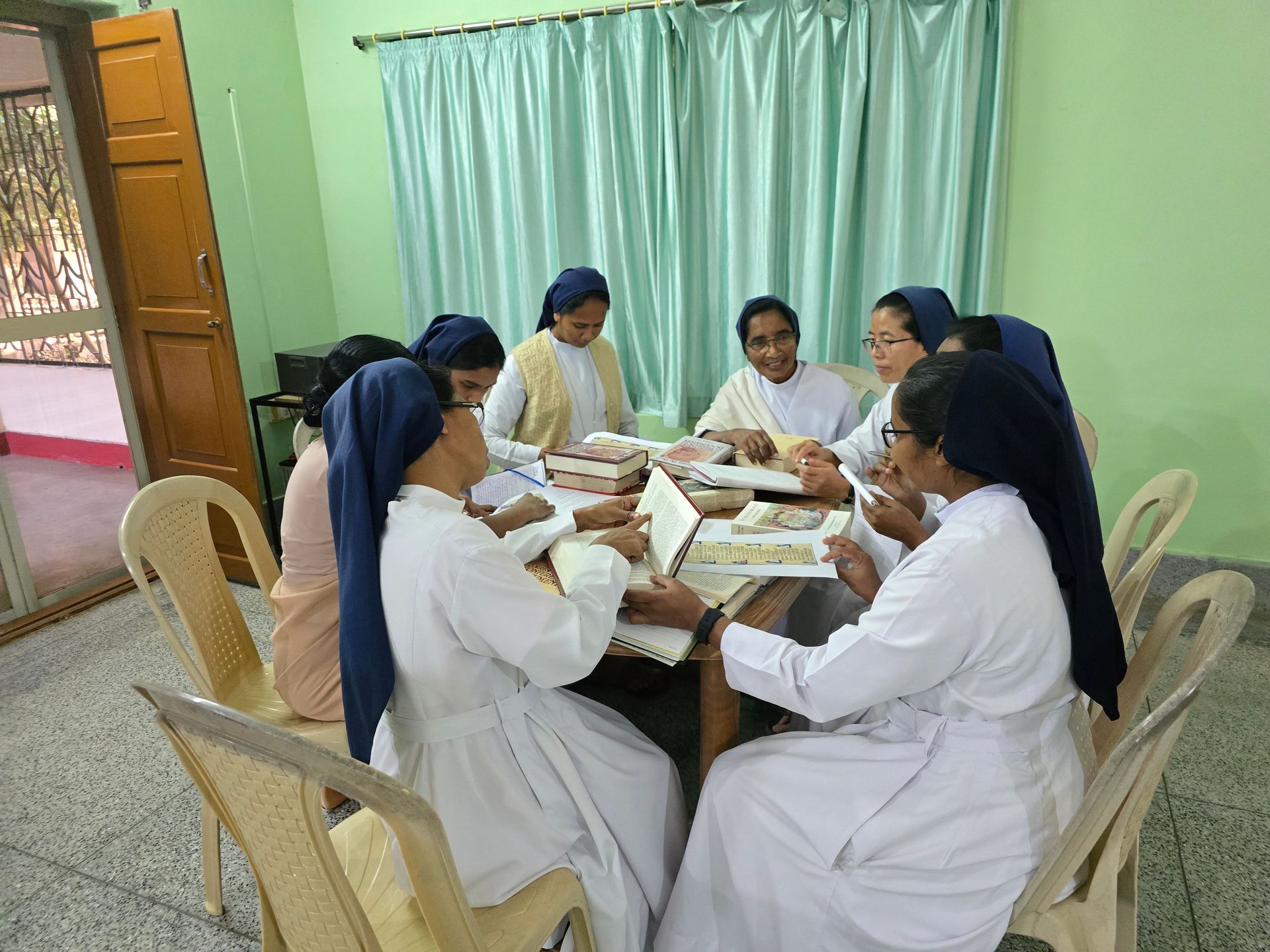Group of nuns in white habits and blue veils sit around a table, studying.