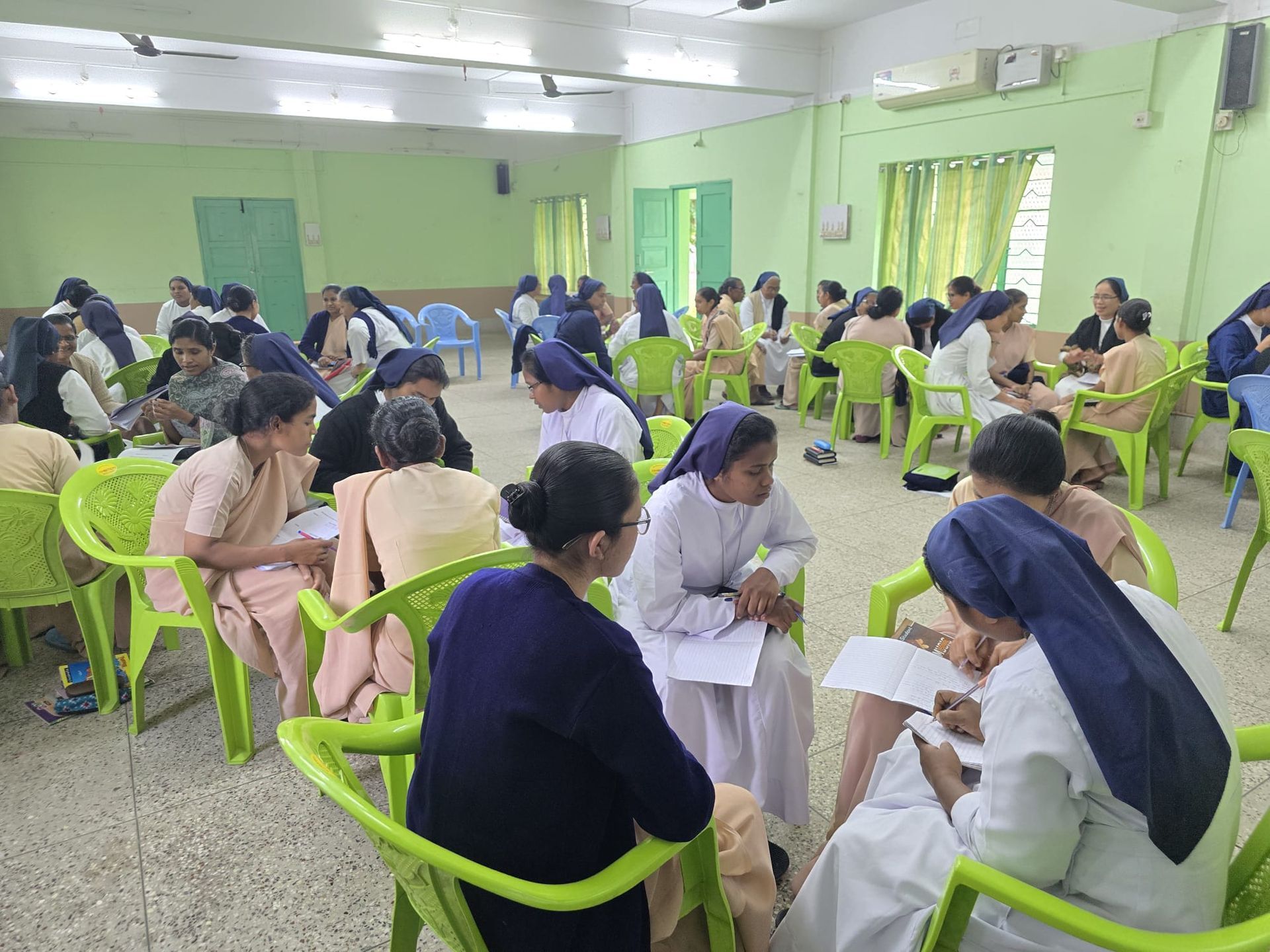 Nuns in a bright room, seated in groups, engaged in discussion; light green chairs and walls.