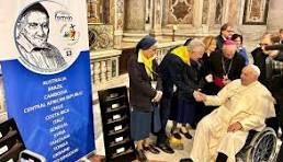 Pope Francis in wheelchair shakes hands with a person. Banner lists countries. Church interior.