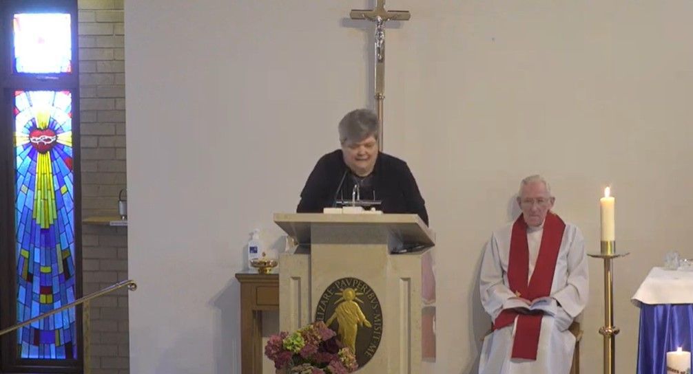 A man is standing at a podium in a church while another man sits in front of a cross.