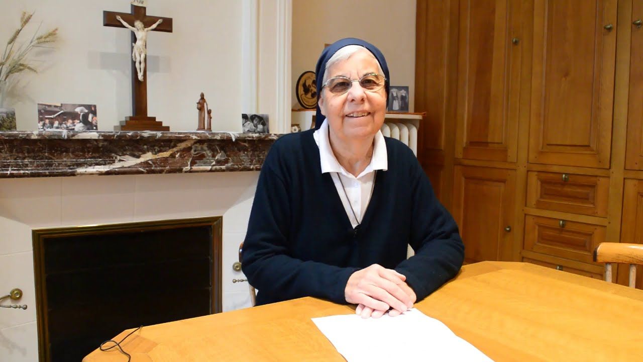 Nun seated at a wooden table, smiling, in a room with a fireplace and crucifix.