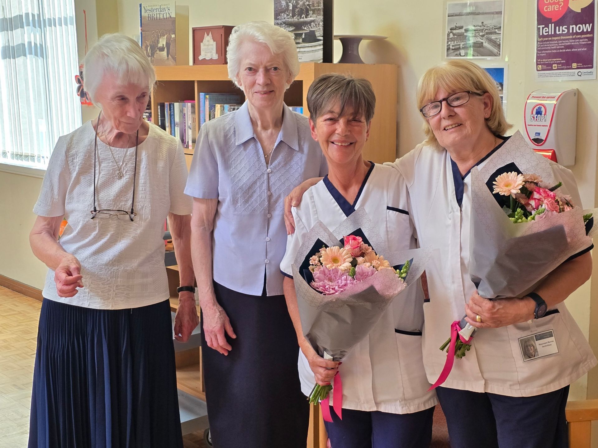 Four women smiling and holding bouquets of flowers, in a library setting. One wears a medical uniform.