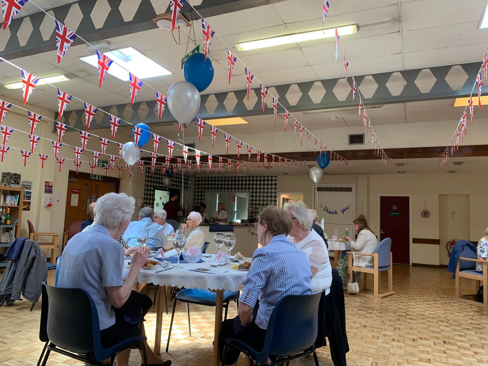 People seated around tables in a decorated room. Union Jack flags and balloons hang from the ceiling.