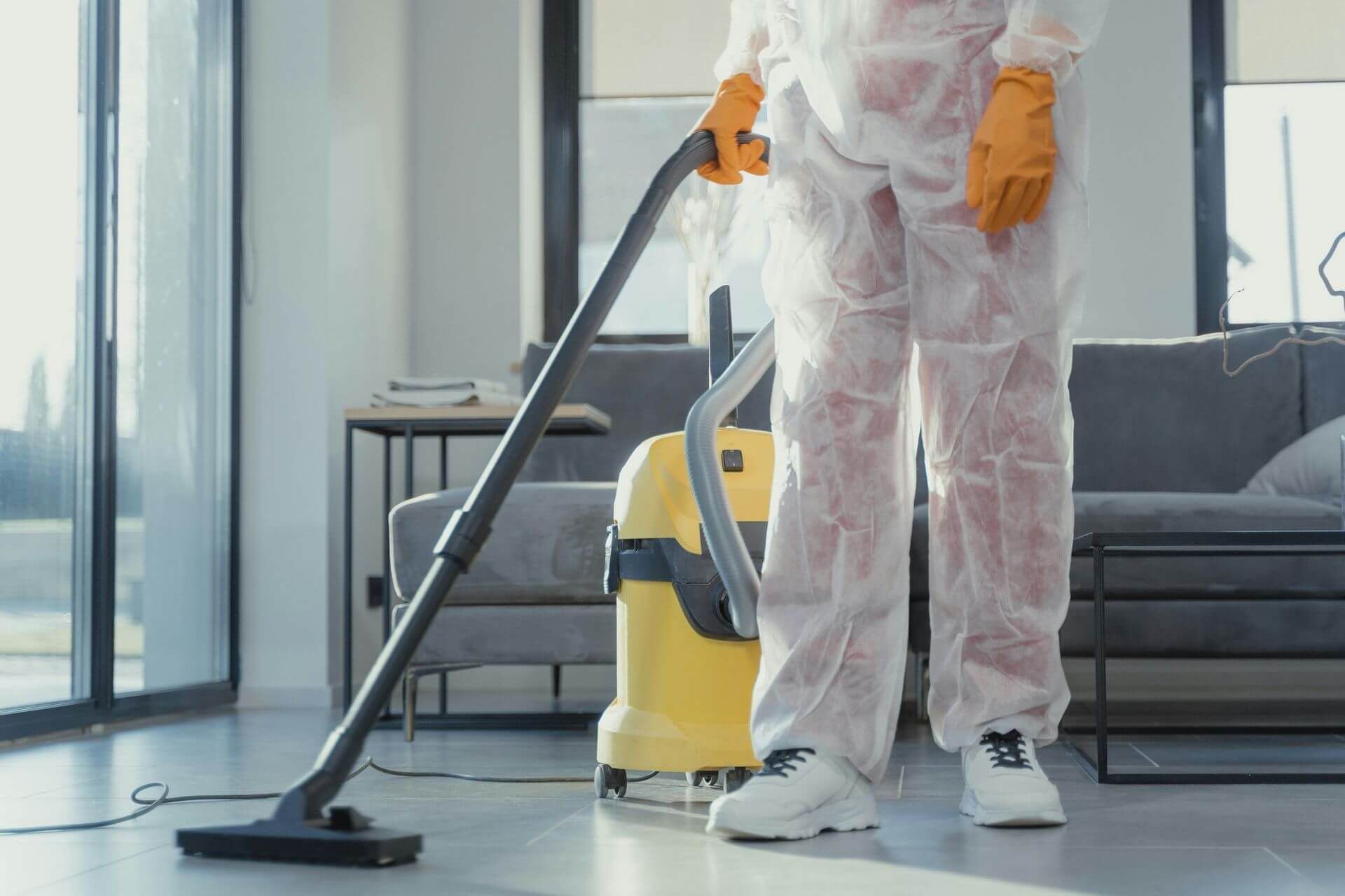 Person cleaning a modern room with a yellow vacuum and mop, wearing white protective coveralls and orange gloves.