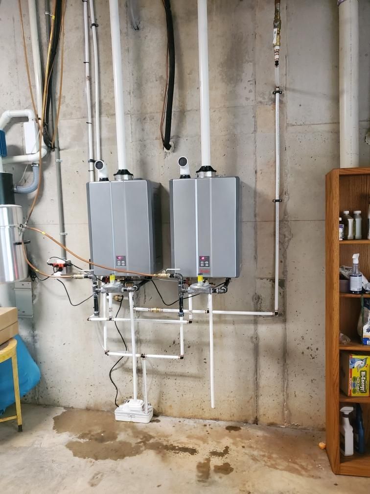 Two wall-mounted gray water heaters with exposed pipes in a utility room with concrete walls.