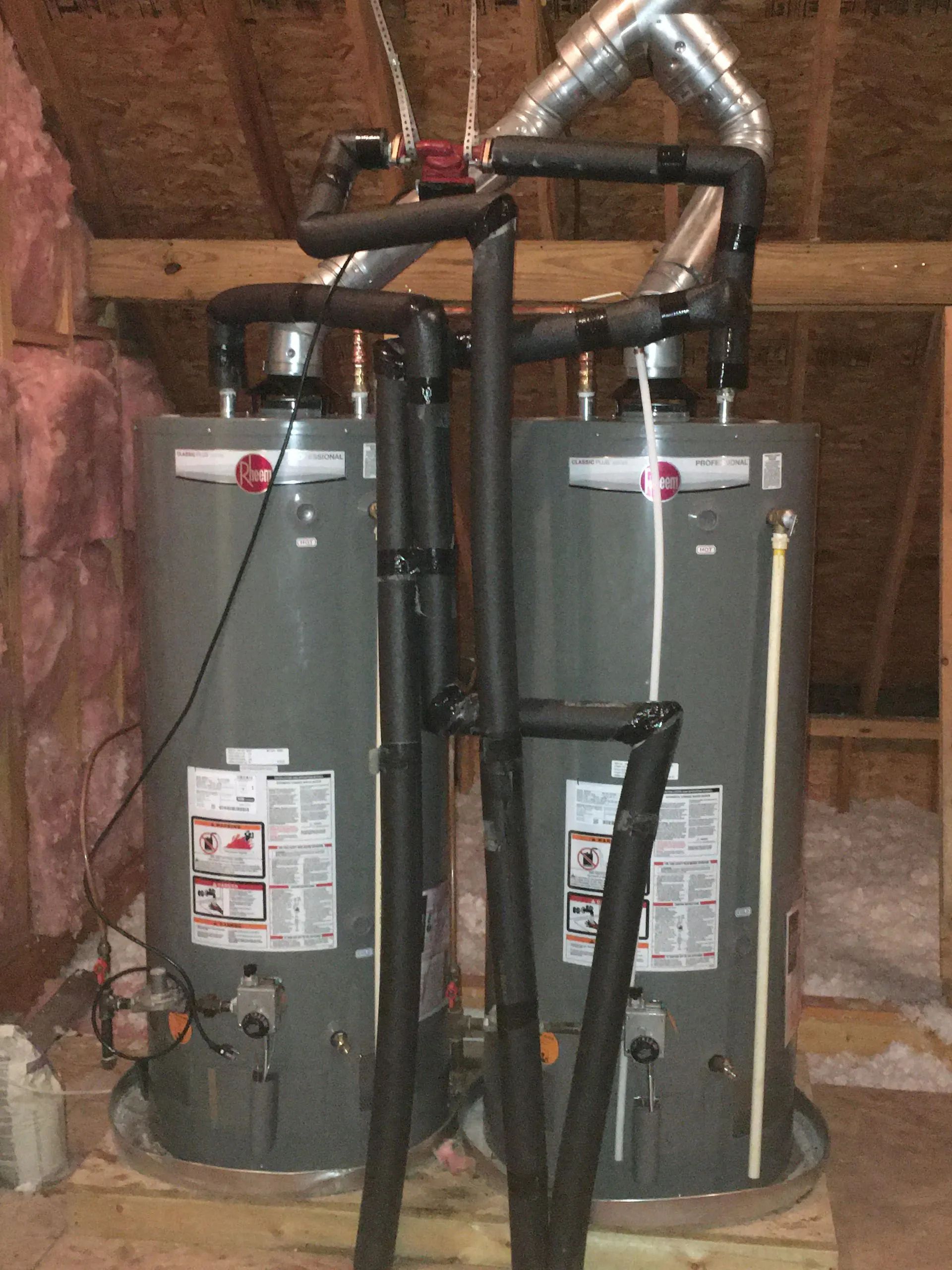 Two gray water heaters with black vent pipes in an attic, side by side among exposed wooden rafters.