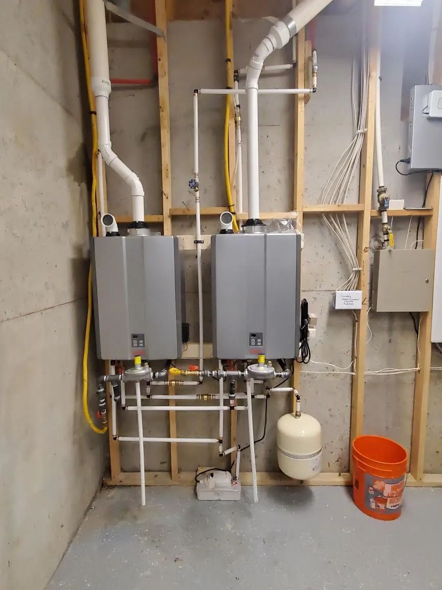 Mechanical room with two gray wall-mounted boilers, white piping, and an orange bucket on a concrete floor.