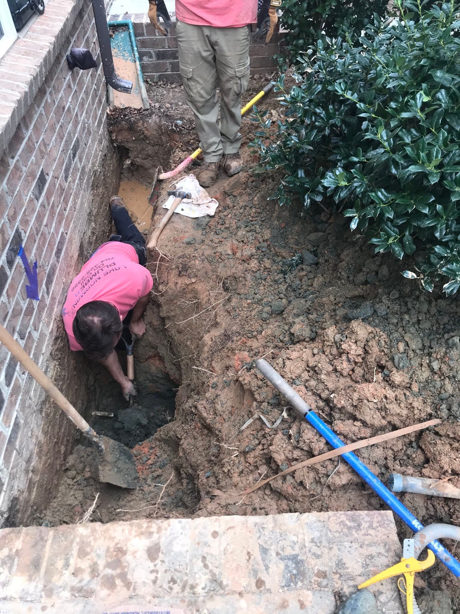 Workers digging a narrow trench beside a brick wall, with tools and hoses scattered along the dirt path.