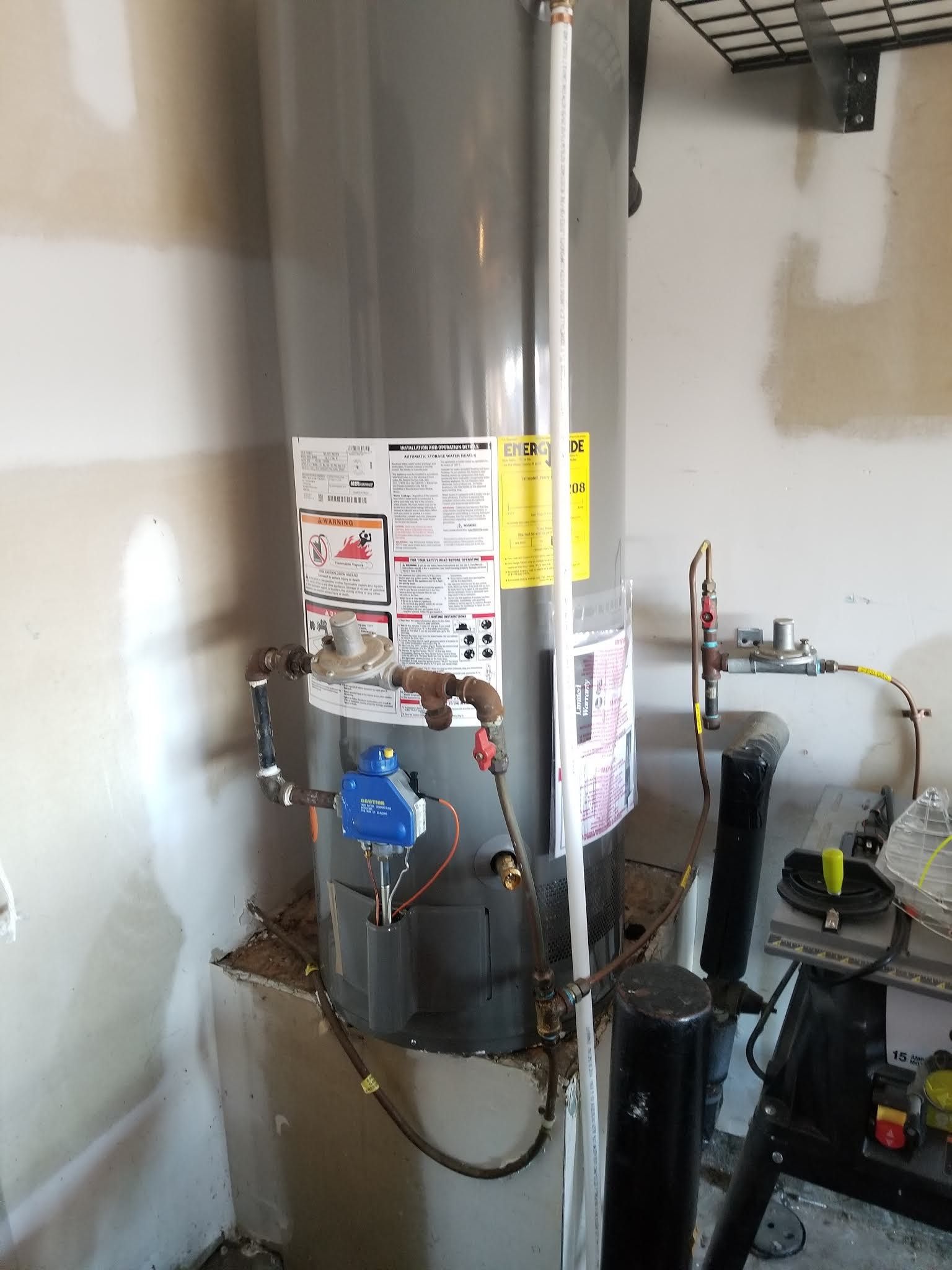 Water heater in a utility room with pipes, gauges, labels, and a gas control panel against unfinished walls.