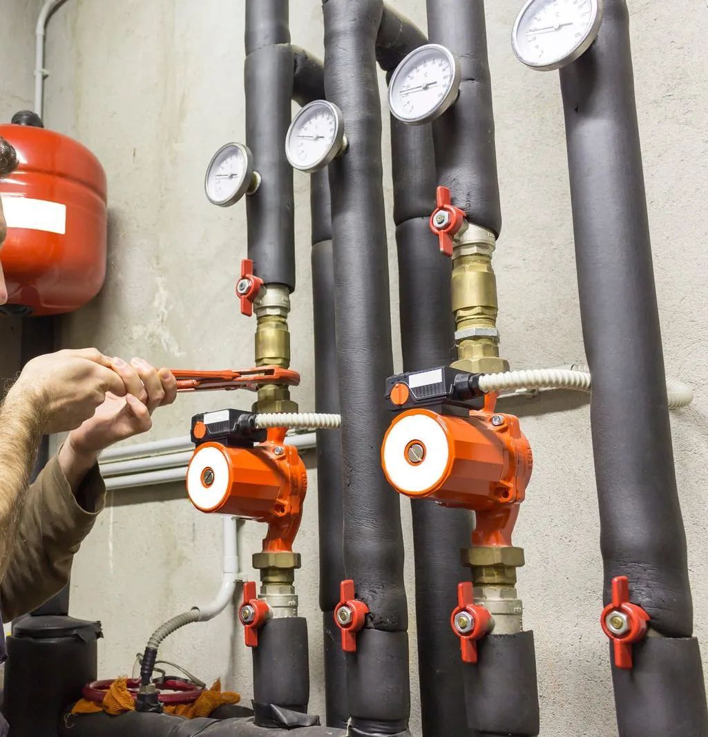 Industrial pumps with pressure gauges and pipes; a hand adjusts an orange valve in a mechanical room.