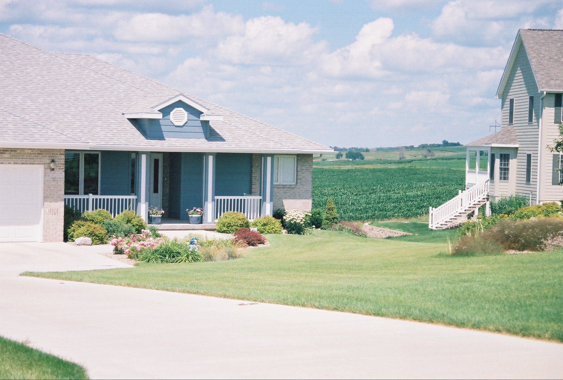 Quiet suburban street with two houses, green lawns, and open fields under a partly cloudy sky