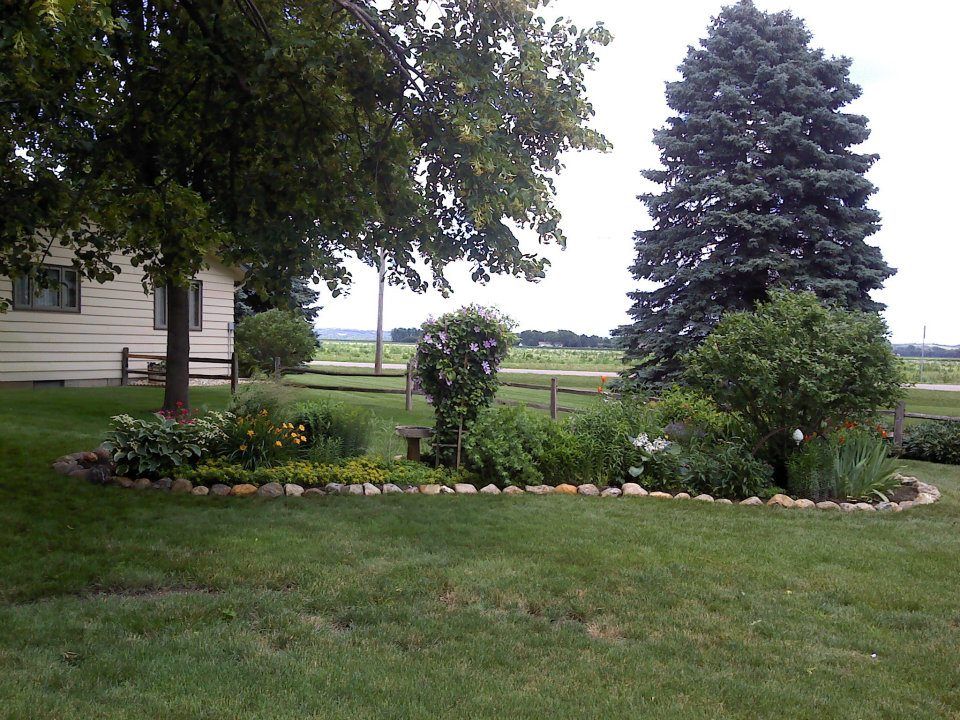 Landscaped garden bed with flowers, shrubs, and trees beside a house on a green lawn.
