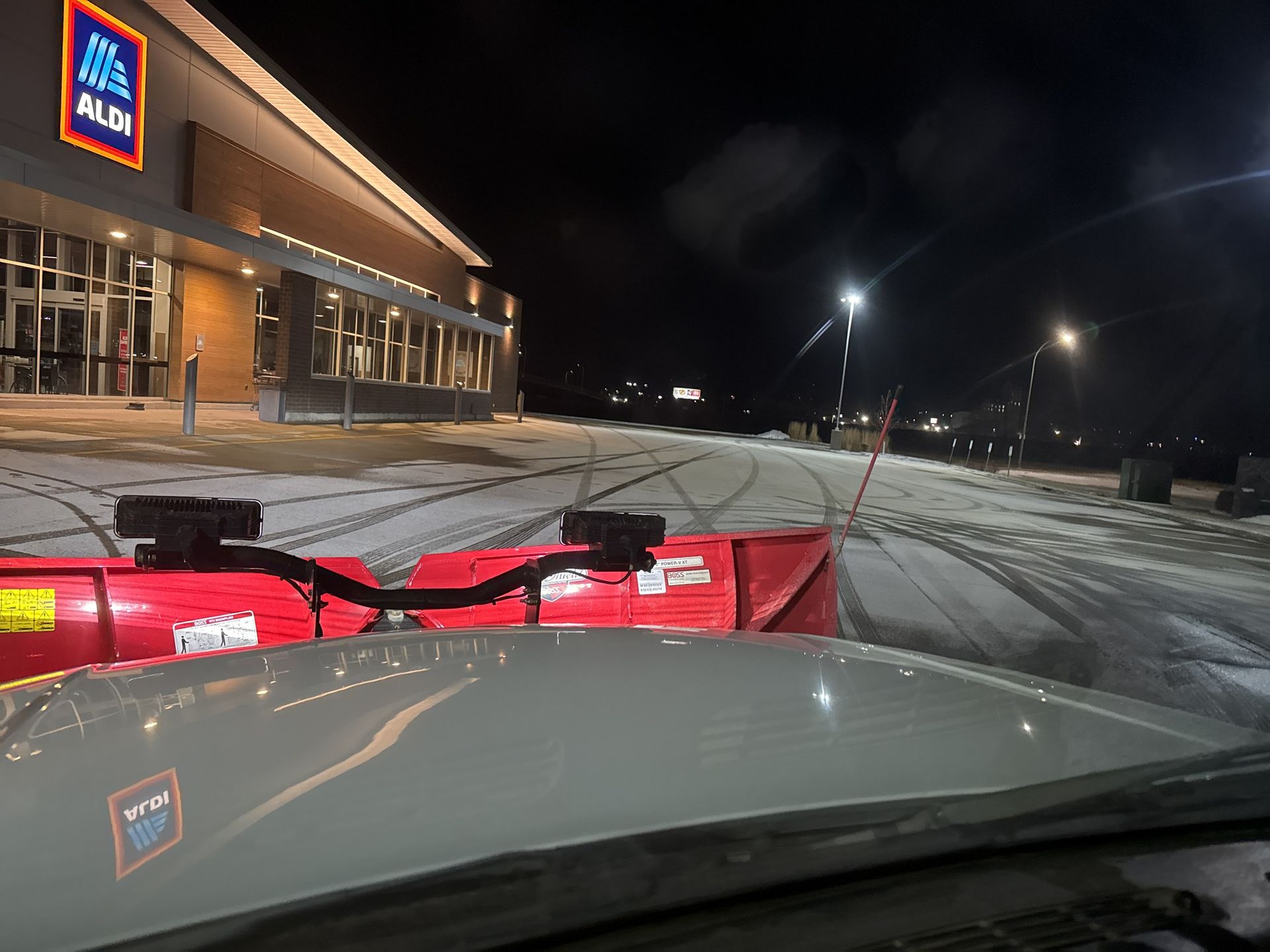 Front-mounted snow plow on a vehicle outside an ALDI store at night in a snowy parking lot