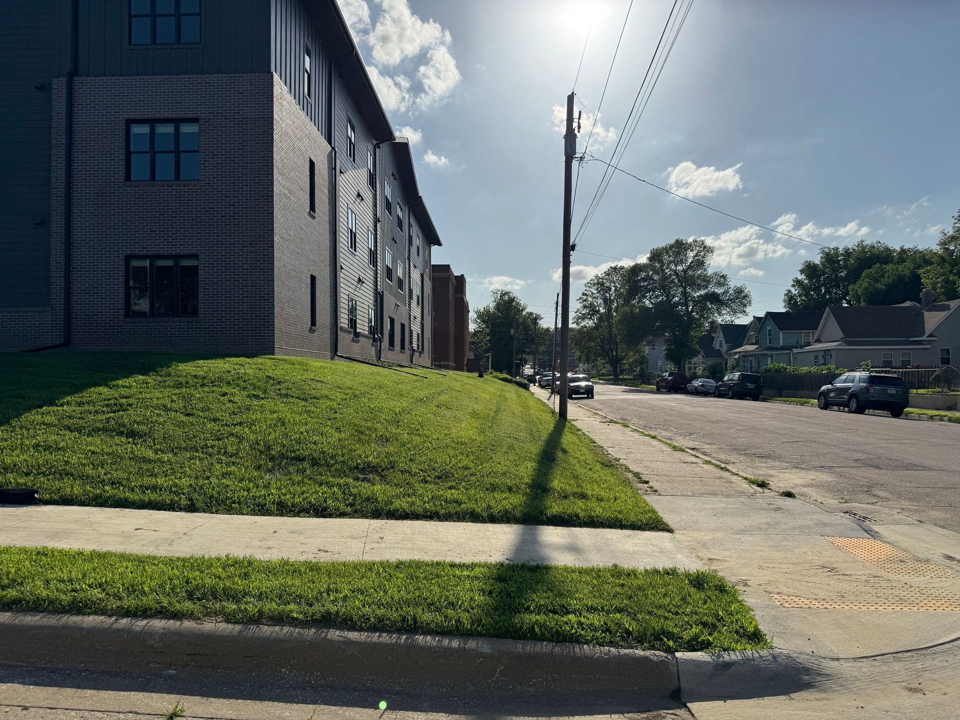 Apartment building beside a sunny residential street with a grassy slope and sidewalk.