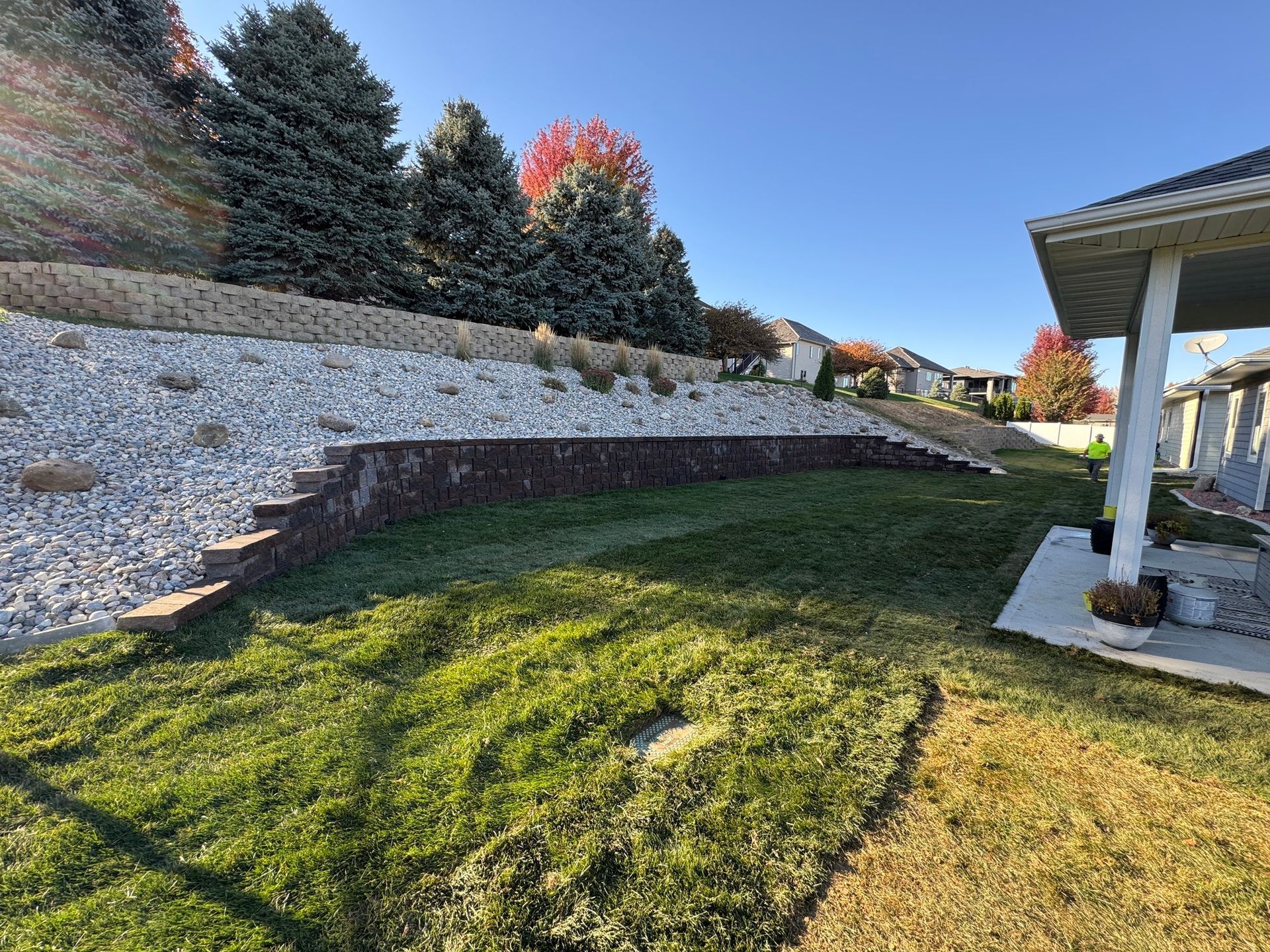 Sloped backyard with green lawn, stone retaining wall, evergreen trees, and a covered patio on the right.