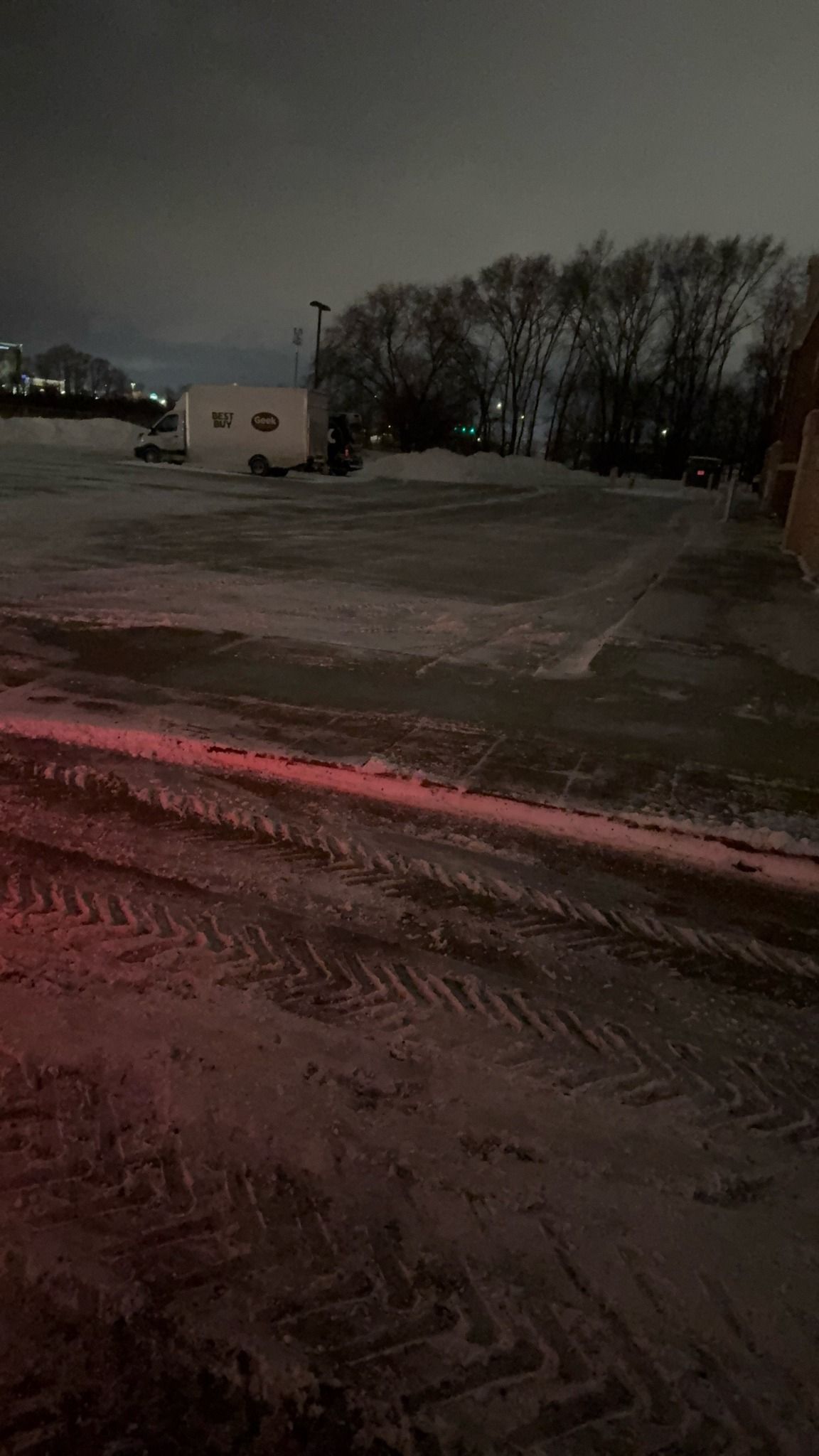 Snowy roadside at night with tire tracks, a building, and bare trees under a dark sky