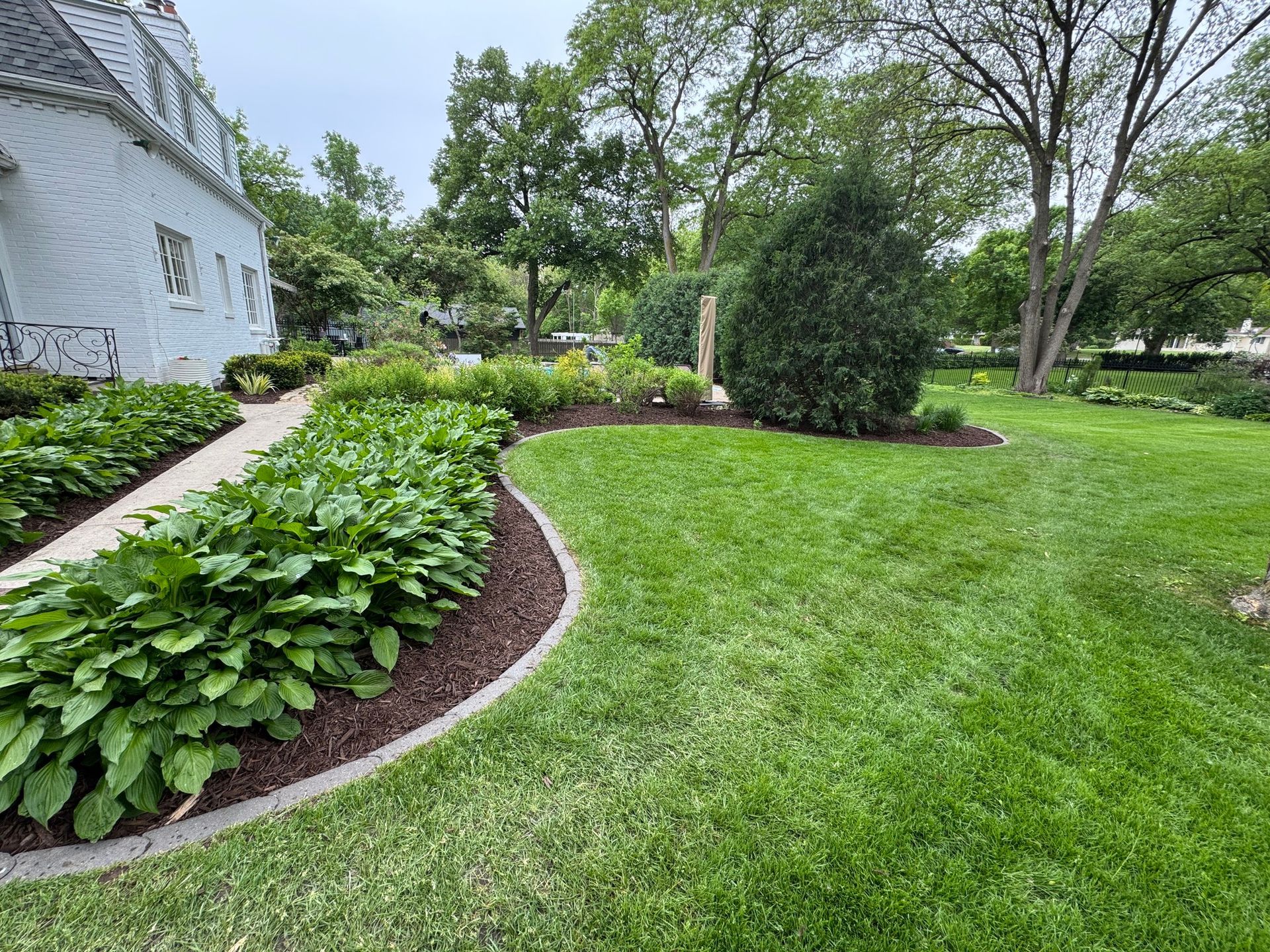Curved garden bed with green shrubs and flowers beside a white house and lawn