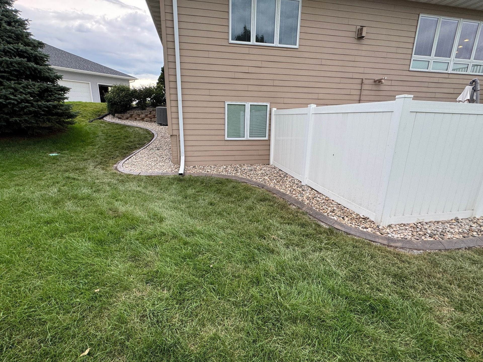 Backyard lawn beside a tan house with a white vinyl fence and stone border