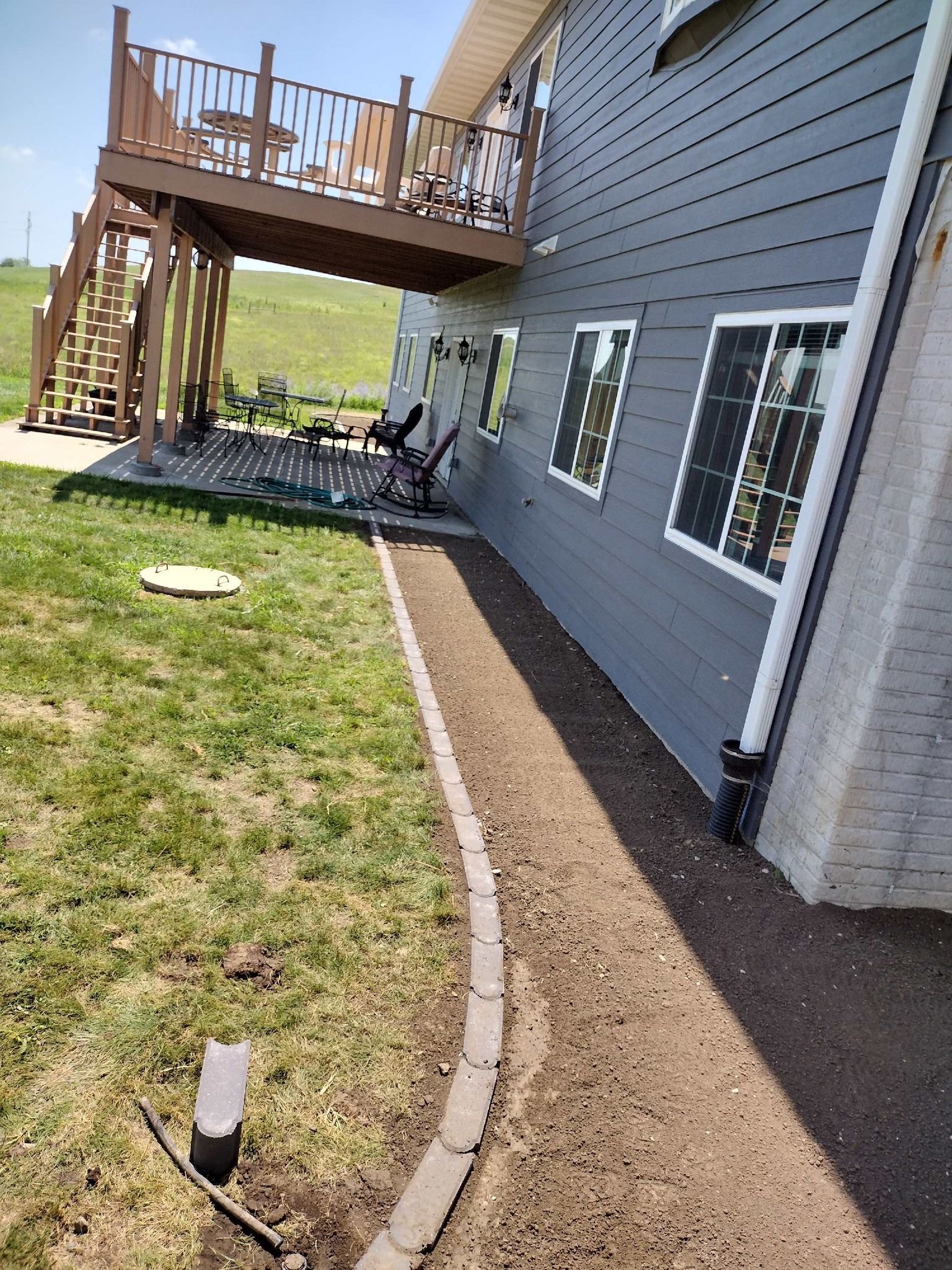Side yard with gray house siding, curved edging, grass, windows, and a raised wooden deck with stairs.