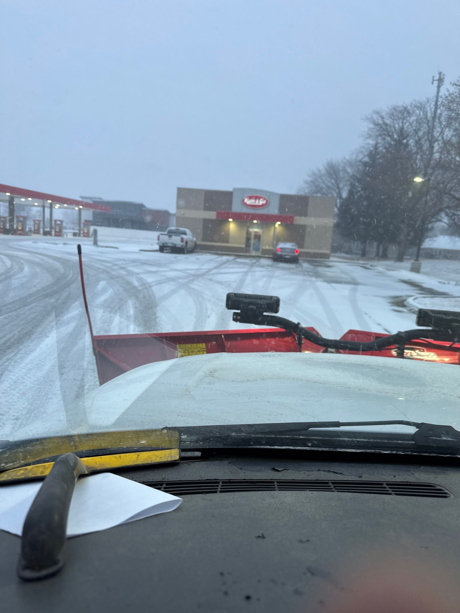 Snowy parking lot seen from inside a truck, with a snow plow blade in front and a building ahead.
