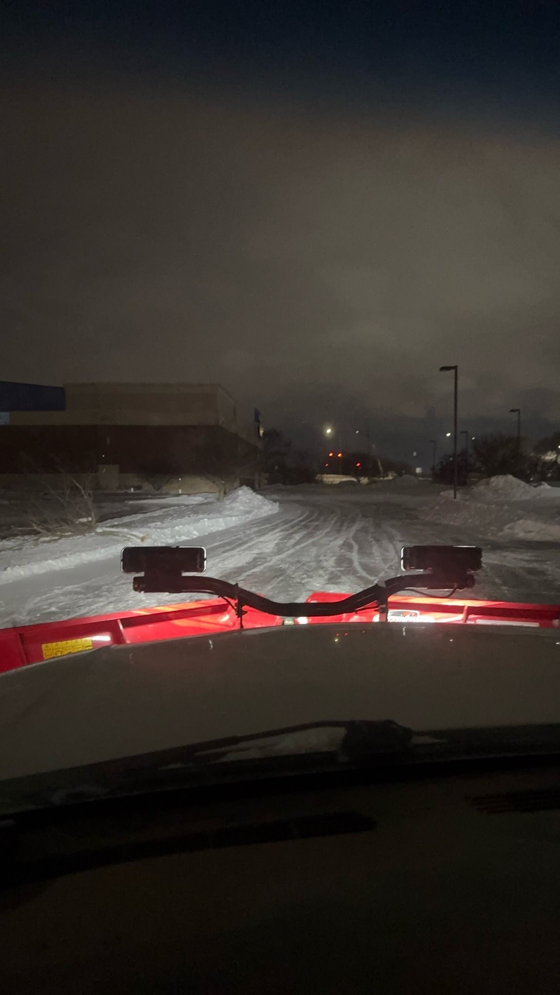 Nighttime snowy road seen from a vehicle, with red hood lights reflecting on the snow-covered street.