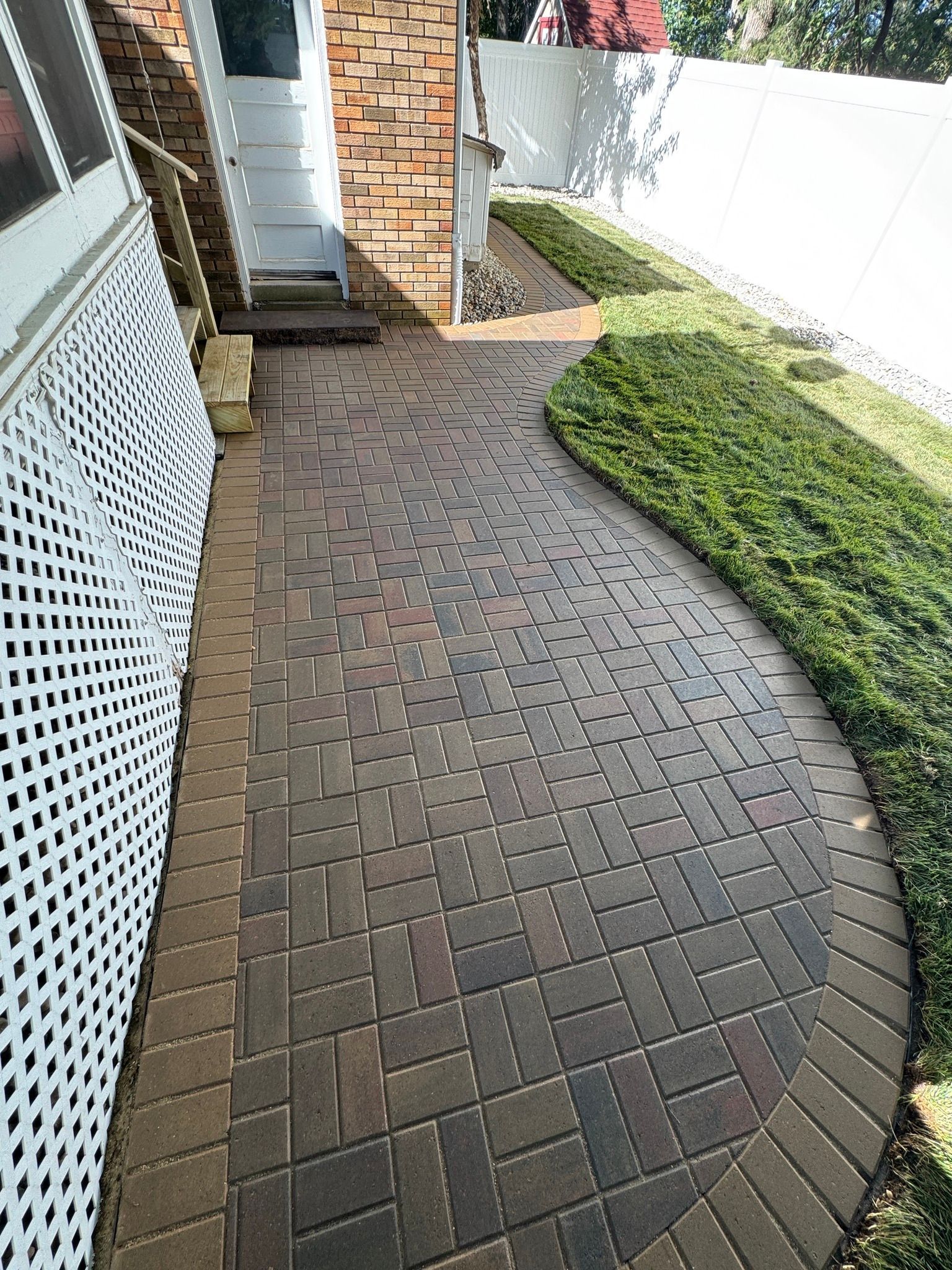 Brick-paved walkway curving beside a house, white fence, and green hedge in a small side yard
