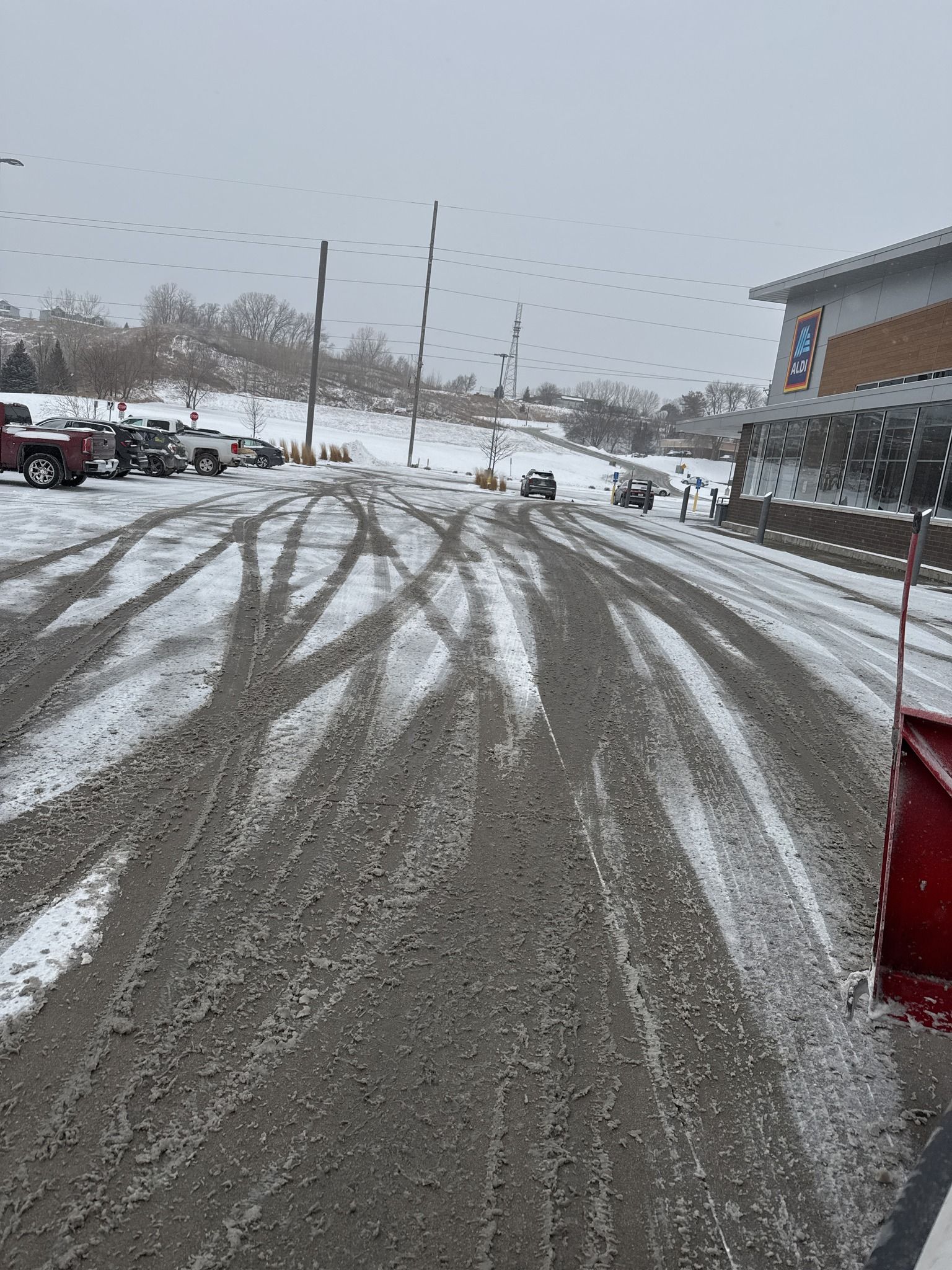 Snow-covered parking lot with tire tracks, parked cars, and a building under a gray sky.