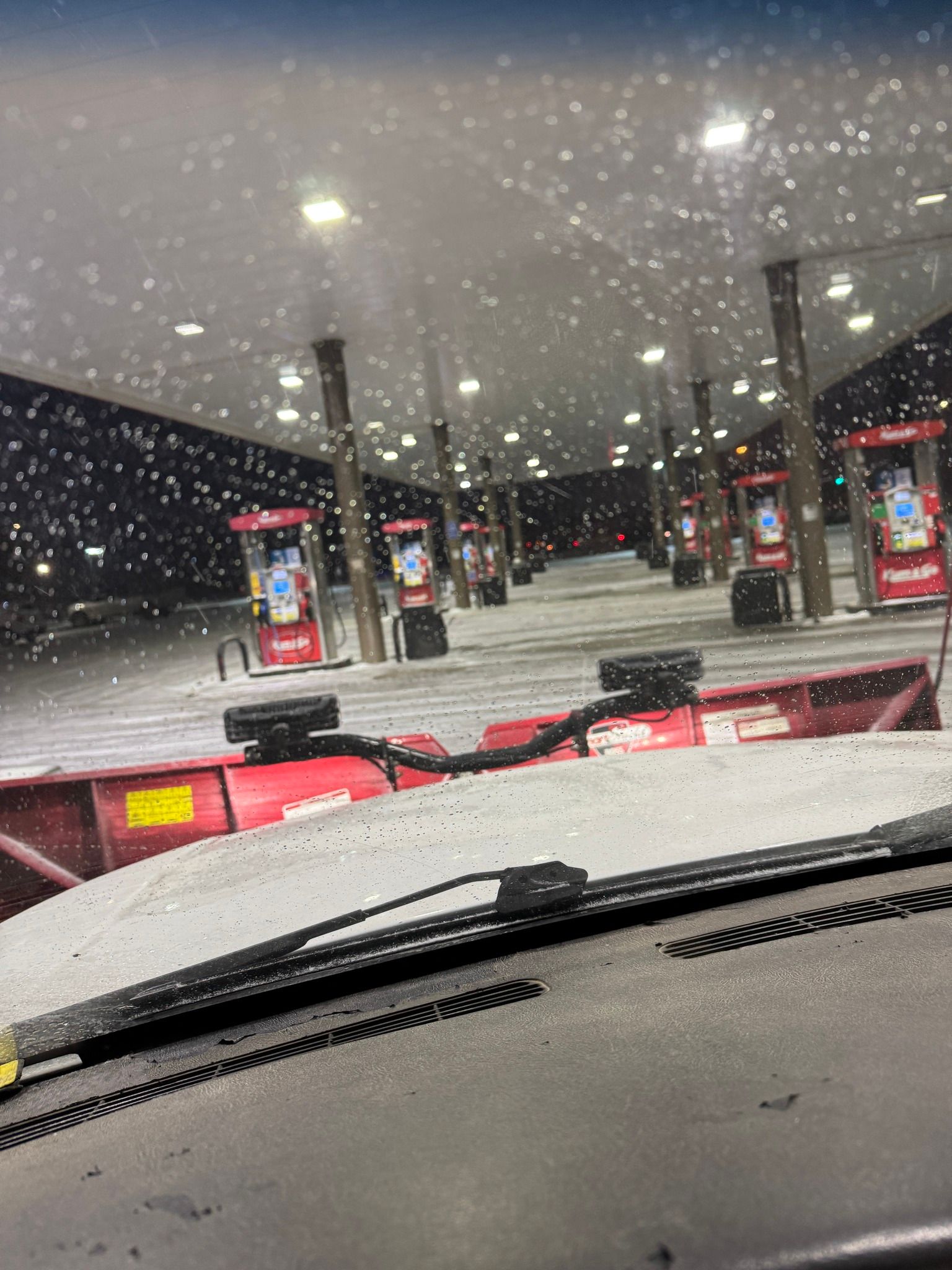 Snowy gas station view through a windshield at night, with bright pumps under a canopy.