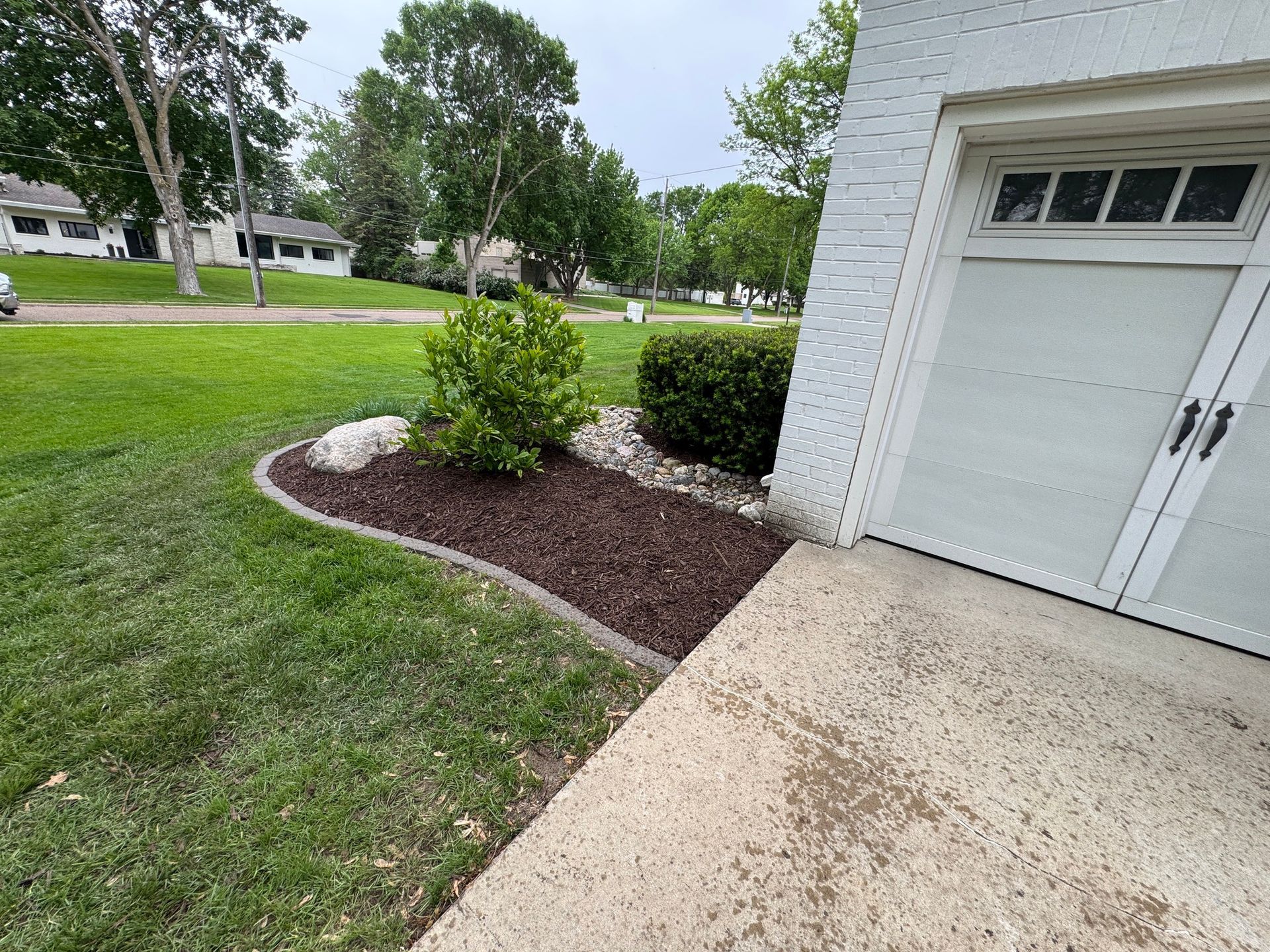 Curved mulch bed with a small shrub beside a garage and driveway on a suburban lawn.