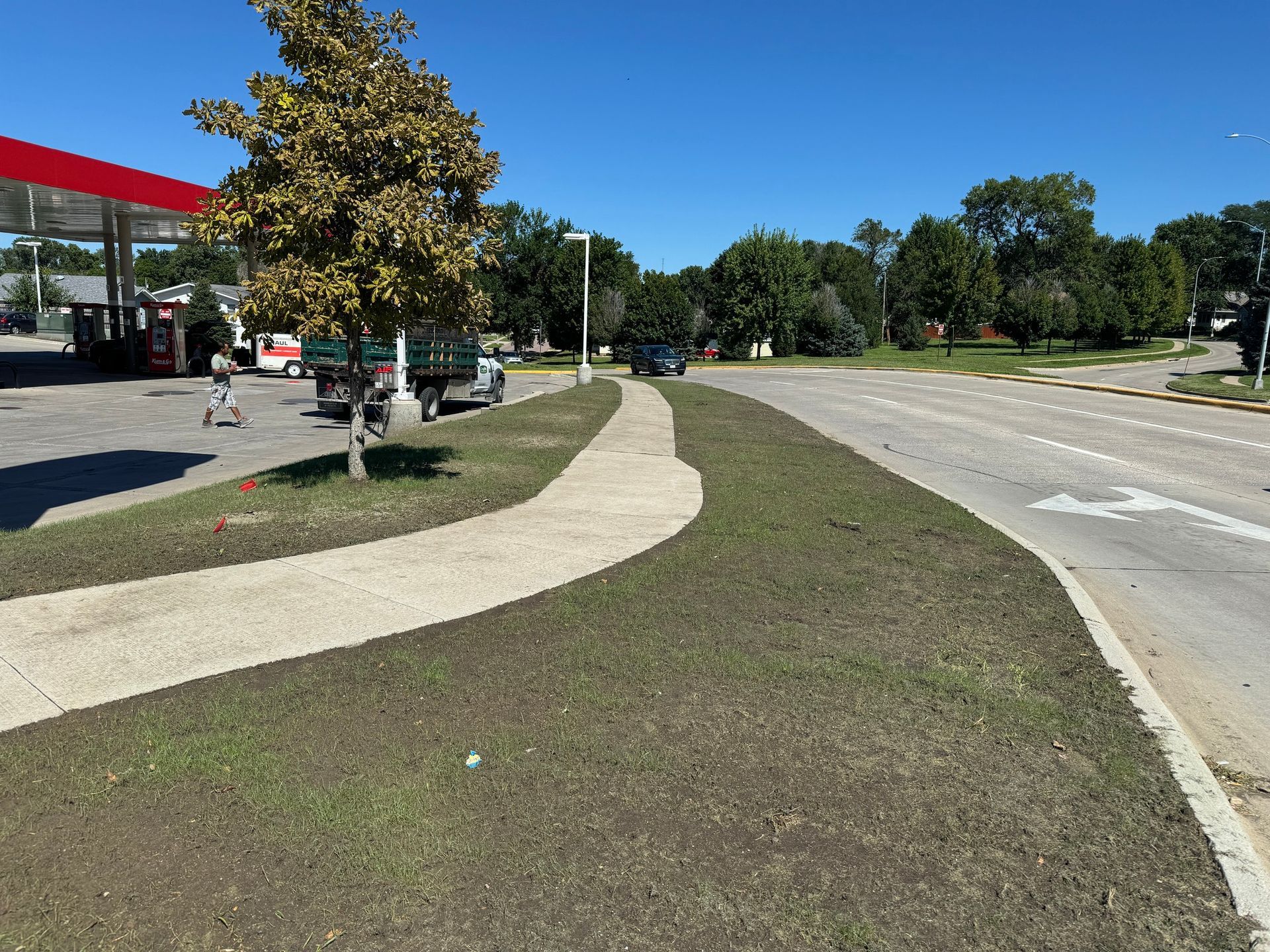 Curved sidewalk beside a road and grassy median near a gas station under clear skies