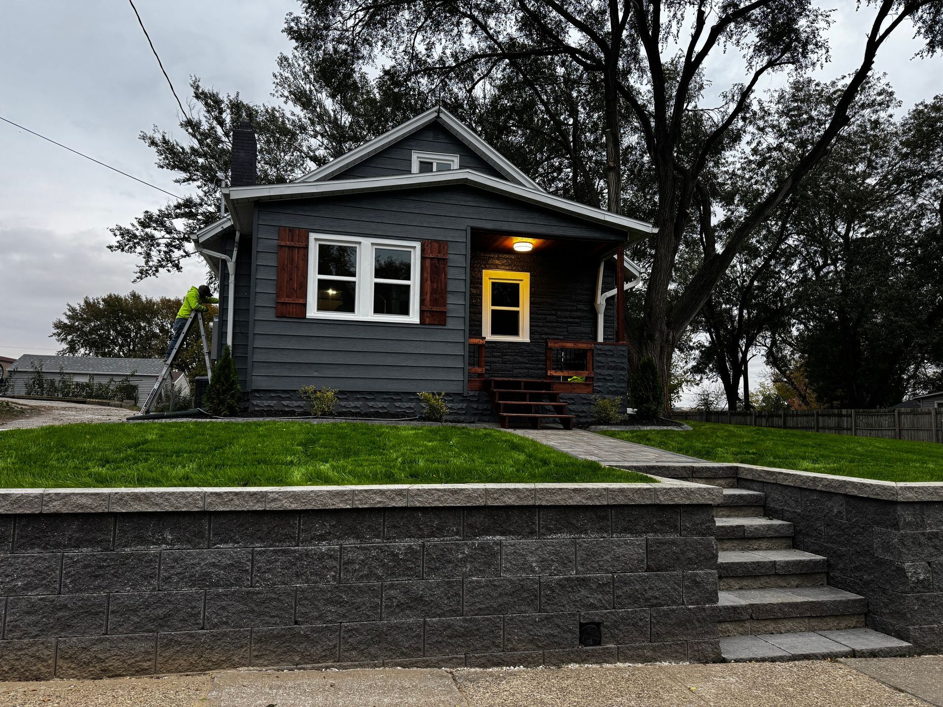 Small dark house with lit porch and stone steps, set on a raised lawn under cloudy skies