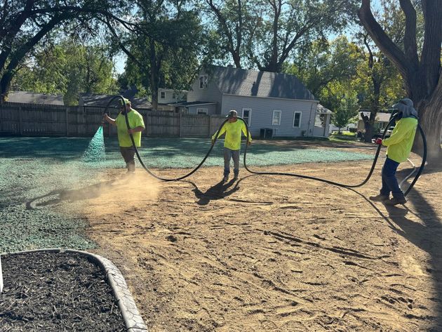 Workers in neon shirts spreading material on a yard under trees, with a house in the background
