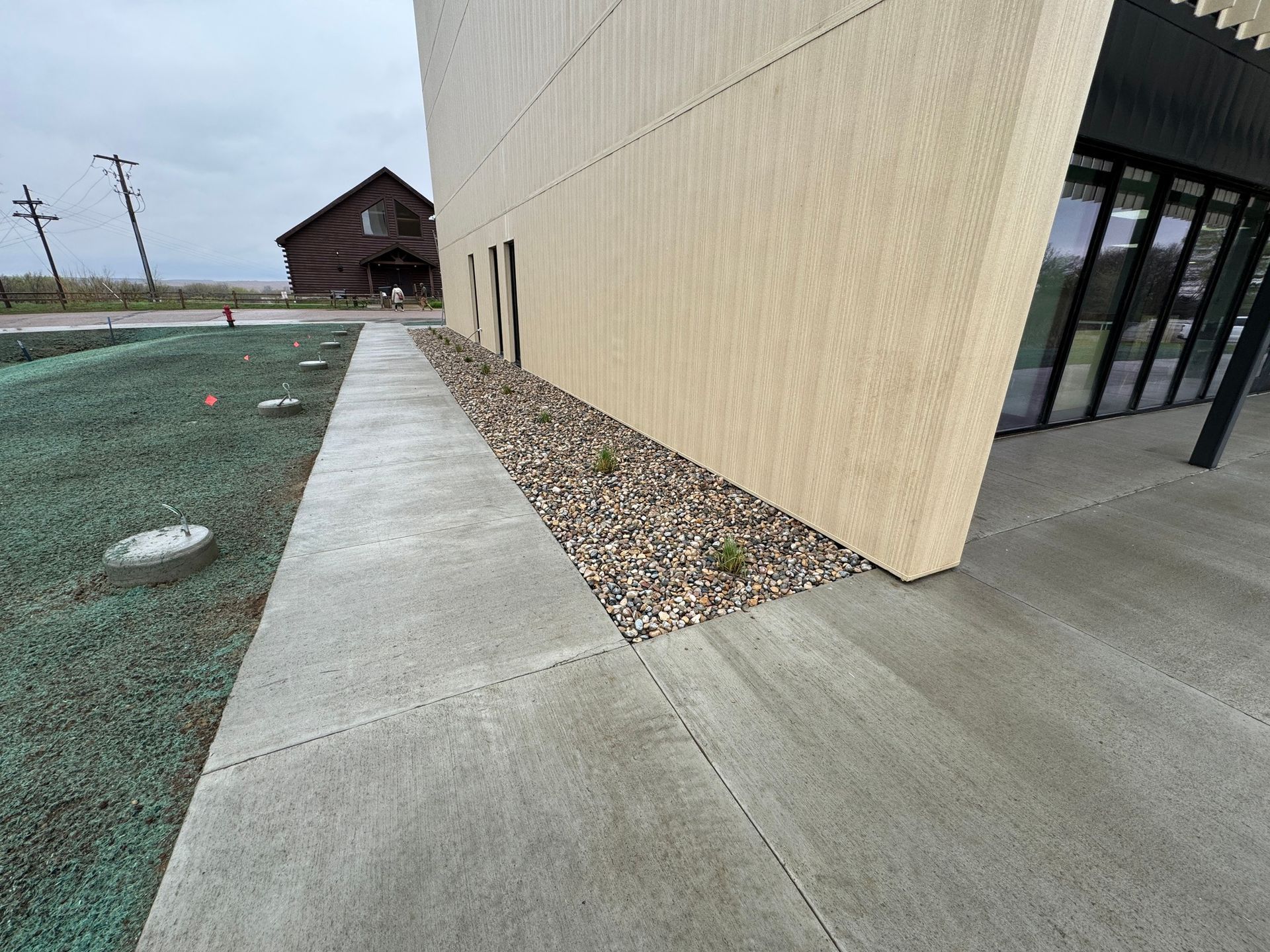 Concrete walkway beside beige building with rock border and green ground cover under overcast sky.