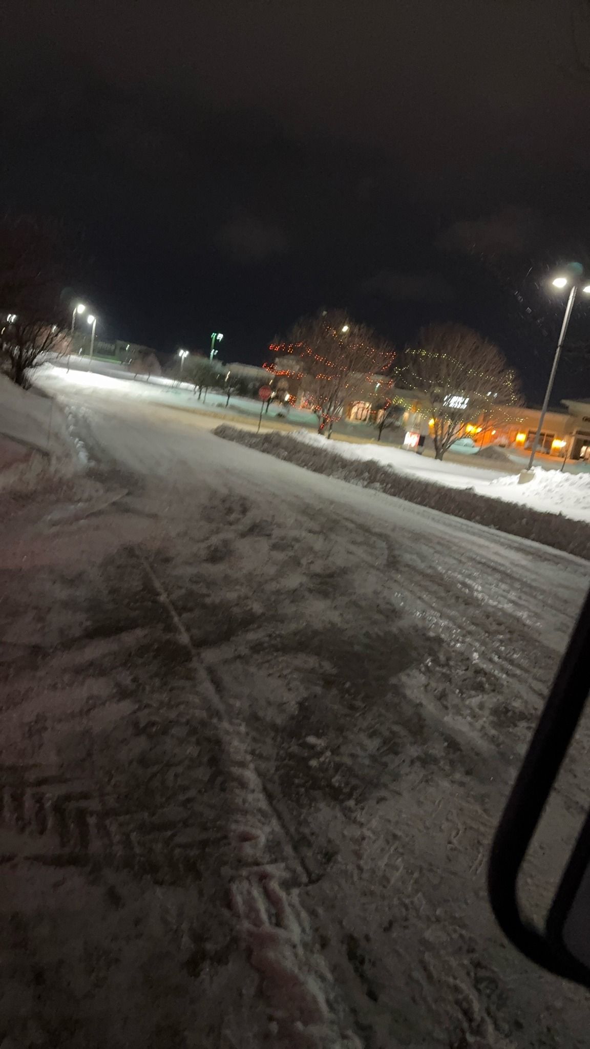 Snow-covered road at night with streetlights and parked cars along the right side.