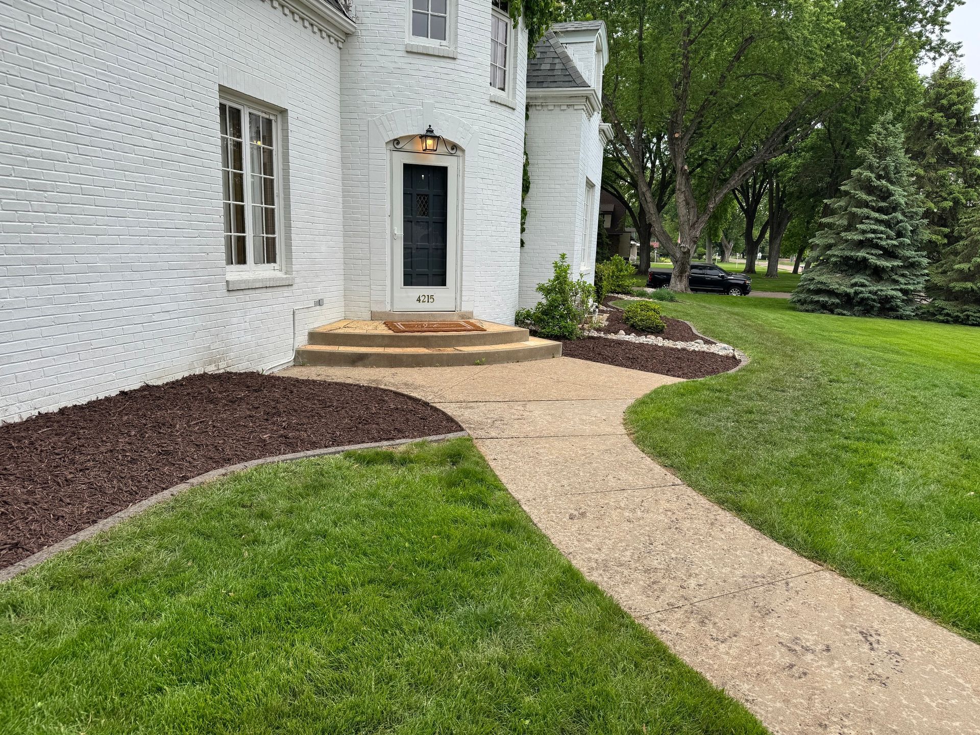 White house entrance with a curved gravel path, green lawn, and mulched flower beds