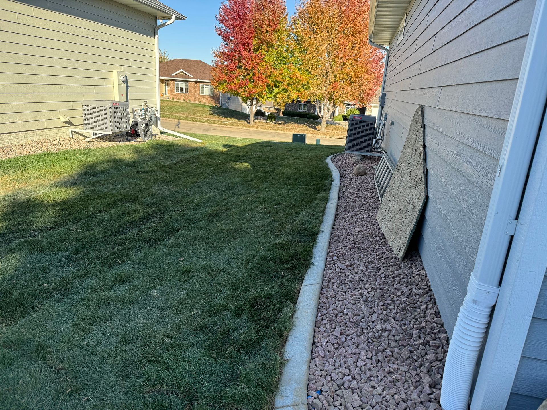 Side yard with grass, white house siding, gravel strip, and autumn trees in the background.