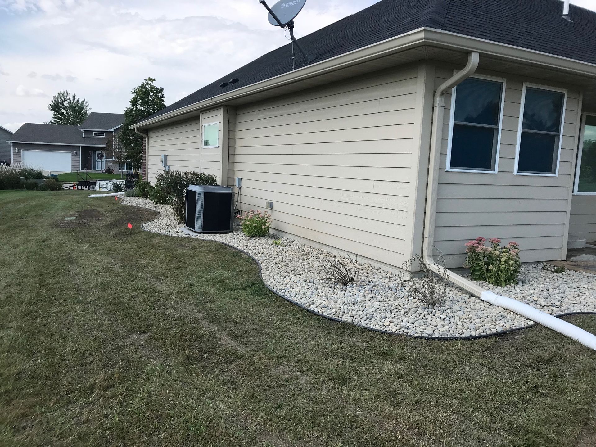 Suburban house exterior with beige siding, gravel border, AC unit, and white downspout beside a grassy yard
