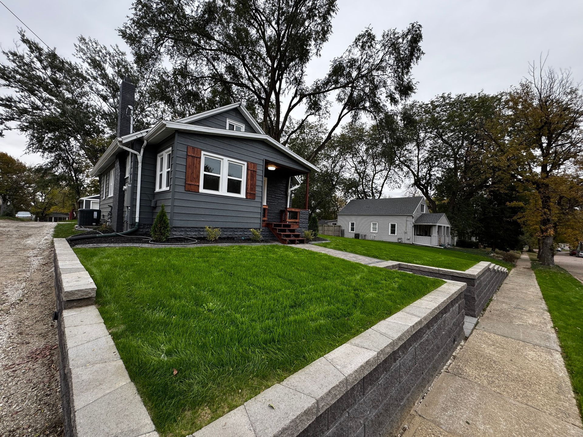 Small gray house with red shutters on a sloped green lawn beside a sidewalk, overcast day